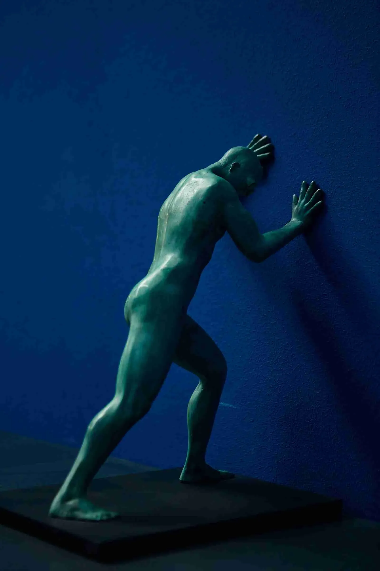 Scultura in bronzo dalla patina verde che raffigura un uomo intento a spingere una parete blu